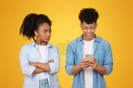 Foto de Milenial mujer afroamericana escépticamente mira a un joven que está felizmente absorto en su teléfono inteligente, ambos con camisas de color azul claro en un alegre fondo amarillo - Imagen libre de derechos