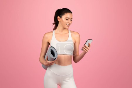 Photo for Smiling millennial caucasian woman in white sportswear hold mat and chatting on phone, isolated on pink studio background. Health care app, weight loss lifestyle, sport and yoga - Royalty Free Image