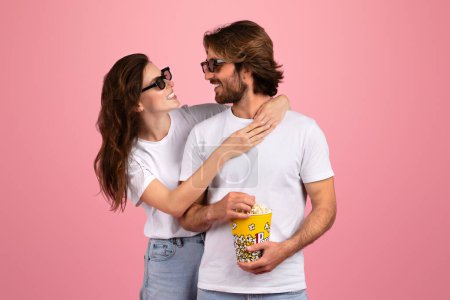 Foto de Una alegre pareja europea en camisetas blancas y gafas de sol compartiendo un momento de risa mientras sostiene un cubo de palomitas de maíz, que representa una cita perfecta para la película sobre un fondo rosa - Imagen libre de derechos