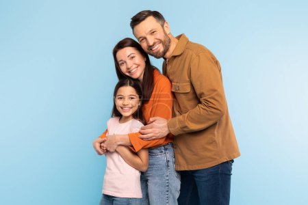 Photo for Family Of Three. Happy Caucasian Man Hugging His Wife And Little Daughter Standing Over Blue Background. Studio Shot Of Cheerful Parents And Kid Girl Posing Together Smiling To Camera - Royalty Free Image