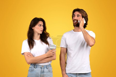 Photo for Skeptical woman holding a coffee cup looking at a smiling man talking on a mobile phone, both dressed in white t-shirts, indicating a disconnect or miscommunication on a yellow background - Royalty Free Image