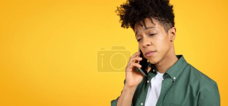 Foto de Enojado joven afroamericano hombre hablando por teléfono celular, mirando el espacio de copia para la publicidad, aislado en el fondo del estudio amarillo, banner web. Concepto de comunicación - Imagen libre de derechos