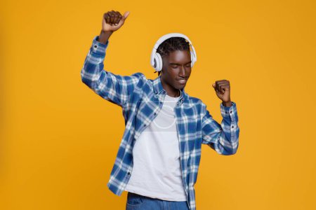 Téléchargez les photos : Jeune homme noir positif appréciant écouter de la musique, danser et s'amuser, homme afro-américain millénaire joyeux portant des écouteurs sans fil posant sur fond de studio jaune, espace de copie - en image libre de droit