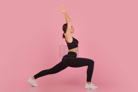 Téléchargez les photos : Fit jeune femme effectuant la pose guerrière dans le yoga sur fond de studio rose, sportive femme millénaire démontrant flexibilité et équilibre, formation en vêtements de sport noirs, vue de côté avec espace de copie - en image libre de droit