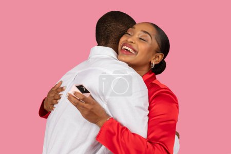 Photo for Overjoyed black woman in red embracing man, showing off her new engagement ring behind his back, moment of pure happiness, pink background - Royalty Free Image