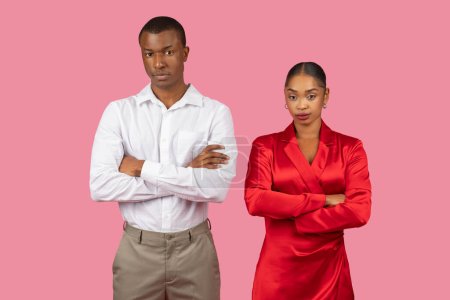 Photo for Serious black man in white shirt and woman in red dress stand with arms crossed, displaying serious expressions and confident stances, pink background - Royalty Free Image