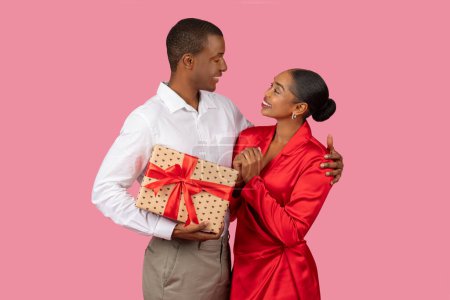 Téléchargez les photos : Aimant couple noir échangeant des cadeaux, avec l'homme donnant cadeau enveloppé à une femme souriante en robe rouge, exprimant affection et bonheur - en image libre de droit