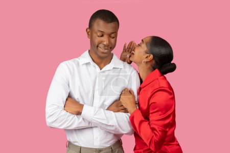 Photo for Black woman in red dress whispers into the ear of smiling man in white shirt, sharing secret or private joke between them, pink background - Royalty Free Image