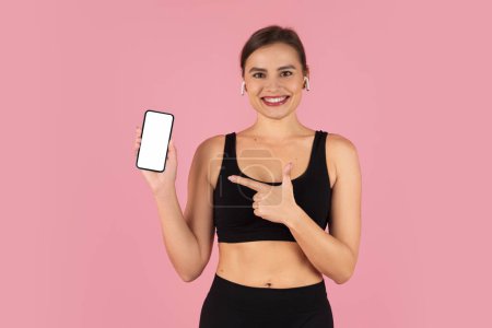Photo for Smiling fitness woman in sportswear joyfully presenting blank smartphone screen, pointing at empty cellphone with one hand, happy young fit lady posing on pink studio background, mockup - Royalty Free Image