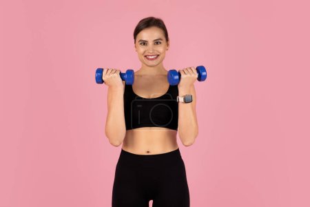 Photo for Smiling sporty woman doing fitness routine, training with blue dumbbells, fit young lady dressed in black activewear exercising against pink studio background, enjoying healthy lifestyle, copy space - Royalty Free Image