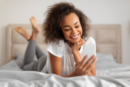 Foto de Sonriendo afroamericana joven mujer mensajes de texto utilizando el teléfono móvil, navegar por Internet y la creación de redes acostado en el estómago en la cama en el interior del dormitorio moderno. Gran aplicación móvil, tecnología - Imagen libre de derechos
