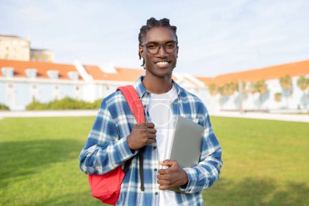 Photo for Education Abroad. Smiling handsome young black guy carrying backpack and laptop standing against backdrop of university campus, looking at camera with confidence. Online learning concept - Royalty Free Image