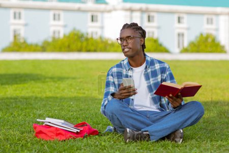 Photo for Young black student guy holding mobile phone and opened book while learning outdoor, young man engaging in e-learning and traditional reading, spending free time outside campus park - Royalty Free Image