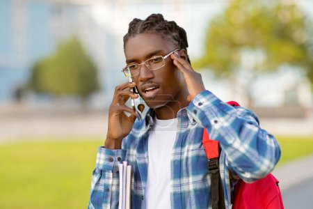 Téléchargez les photos : Portrait de jeune homme noir en lunettes tient sac à dos et parler par smartphone sur le campus en plein air, discuter du problème au téléphone, faire face à des défis dans les activités universitaires - en image libre de droit