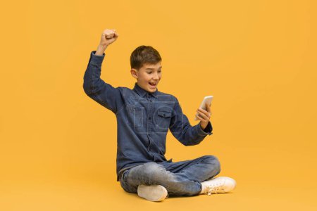 Téléchargez les photos : Joyeux garçon adolescent regardant l'écran du smartphone et célébrant le succès tout en étant assis sur le sol en studio, heureux enfant mâle levant le bras dans la pose de la victoire, acclamant de bonnes nouvelles sur fond jaune - en image libre de droit