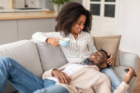 Foto de Cónyuges negros felices tomando café relajándose juntos en el sofá, viendo la televisión en la cómoda sala de estar, hombre descansando de cabeza en las piernas de las esposas, disfrutando de un fin de semana perezoso en interiores - Imagen libre de derechos