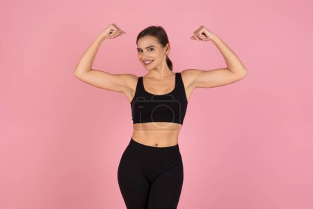 Foto de Mujer fitness segura y sonriente en ropa deportiva flexionando sus bíceps y mostrando su fuerza física, atlética joven hembra de pie sobre fondo de estudio rosa suave, espacio de copia - Imagen libre de derechos