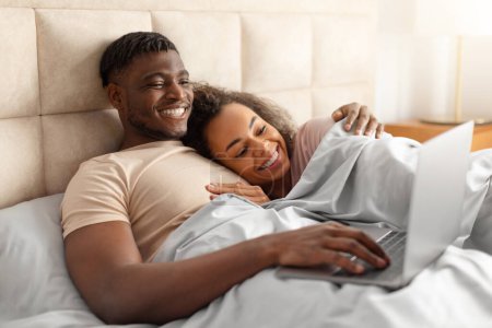 Téléchargez les photos : Joyeux couple afro-américain regardant un film sur ordinateur portable couché dans le lit à la maison. Jeunes conjoints bénéficiant de la technologie Internet et week-end en famille Matin, Websurf Via Gadget - en image libre de droit