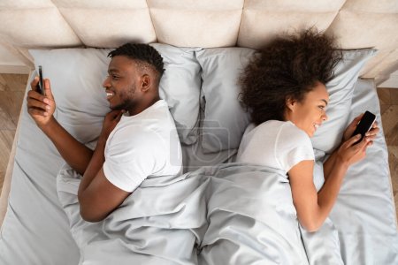 Téléchargez les photos : Tromper le mari et la femme noirs en s'amusant à utiliser des téléphones intelligents tout en étant couché dans le lit dos à dos, passer du temps au lit en ligne dans la chambre moderne. Vue du dessus Prise de vue de couple textos et websurf - en image libre de droit
