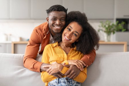 Téléchargez les photos : Bonheur et amour. Joyeux mari afro-américain étreignant la femme de dos tandis que la femme assise sur le canapé, souriant à la caméra posant pour le portrait de famille dans le salon moderne intérieur - en image libre de droit