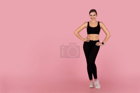 Téléchargez les photos : Jeune femme confiante posant en vêtements de sport sur fond de studio rose, belle femme millénaire présentant un mode de vie sain, debout avec les mains sur la taille et souriant à la caméra, pleine longueur - en image libre de droit