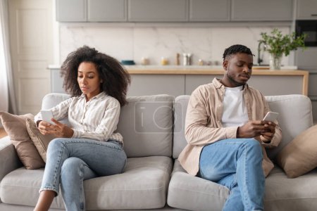 Téléchargez les photos : Phubbing. Mécontent couple afro-américain ennuyé utilisant des smartphones assis sur différents côtés du canapé, ignorant conjoint, ayant un problème dans la relation, accordant plus d'attention aux gadgets à la maison - en image libre de droit