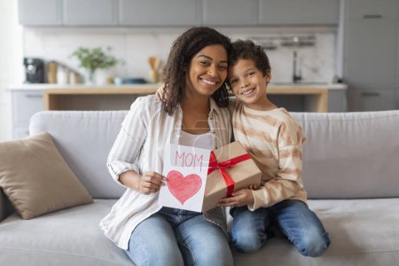 Photo for Mothers Day Gift. Happy black little boy greeting mom with handmade card and wrapped present box, cheerful family of two sitting on couch at home and smiling at camera, mommy and male kid embracing - Royalty Free Image