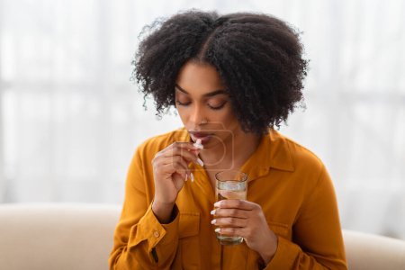 Téléchargez les photos : Triste jeune femme afro-américaine prendre des pilules et boire un verre d'eau dans le salon intérieur. Traitement, soins de santé à domicile, analgésiques pour maux de tête, problèmes et maladies, vitamines, antidépresseurs - en image libre de droit