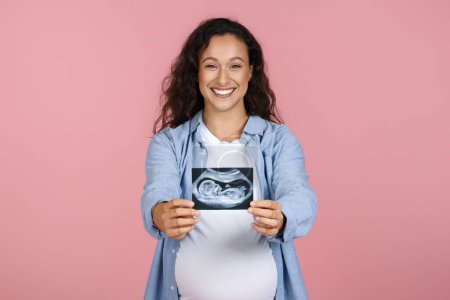 Photo for Happy beautiful young pregnant woman wearing casual clothing showing sonogram ultrasound picture of her baby and smiling, posing isolated on pink studio background - Royalty Free Image