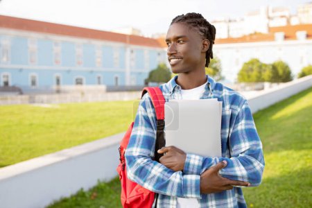 Photo for Black college student guy hugs his laptop standing with backpack near university building outdoor, portrait of young man in casual posing with computer looking aside, ready for education abroad - Royalty Free Image