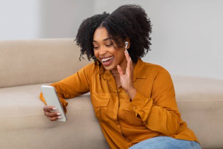 Foto de Sonriente joven rizado señora afroamericana charlando por teléfono, saludando de la mano, tiene videollamada en el interior de la sala de estar. Gadget para reuniones en casa, comunicación remota y aplicación - Imagen libre de derechos