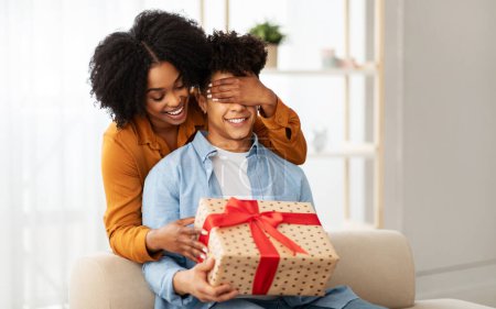 Photo for A joyful african american woman playfully covers a mans eyes from behind, both smiling with anticipation, as they hold a large, heart-patterned gift box with a red ribbon - Royalty Free Image