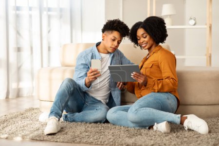 Téléchargez les photos : Habillé occasionnellement en denim, un jeune couple afro-américain est assis sur un tapis moelleux, absorbé dans leurs appareils, avec l'homme tenant un téléphone et la femme une tablette dans un cadre serein à la maison - en image libre de droit
