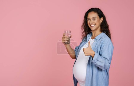 Foto de Mujer joven embarazada sosteniendo un vaso de agua sobre fondo rosa, mostrando el pulgar hacia arriba y sonriendo a la cámara, el concepto de la cantidad de agua consumida, espacio de copia - Imagen libre de derechos