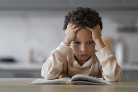 Téléchargez les photos : Stressé petit noir garçon lecture livre à la maison, écrasé preteen afro-américain mâle assis au bureau et tenant la tête dans les mains, faire des devoirs, souffrir des difficultés d'apprentissage, gros plan - en image libre de droit