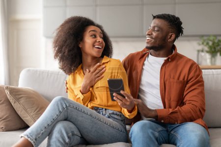 Photo for Young african american spouses using cell phone together and having fun online, scrolling through apps and websites, laughing while sharing internet finds, sitting together on sofa at home - Royalty Free Image