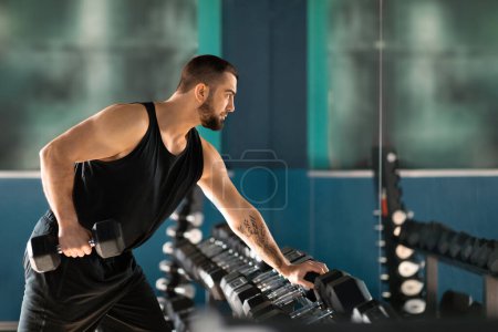 Photo for Masculine young man with beard and tattoos lifting heavy dumbbells, handsome athletic male getting ready for weightlifting in gym, sporty guy training in modern sport club interior, side view - Royalty Free Image
