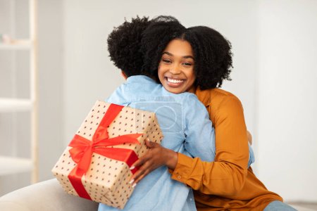 Foto de Una mujer afroamericana con una sonrisa radiante da un cálido abrazo a una persona mientras sostiene una caja de regalo con dibujos del corazón con una cinta roja, que simboliza el afecto y la alegría - Imagen libre de derechos