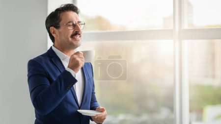 Téléchargez les photos : Homme d'affaires mature caucasien positif en costume bénéficie d'une tasse de café et de détente dans la fenêtre intérieure moderne du bureau, panorama. Travail le matin, affaires, création d'idées, pause et repos - en image libre de droit