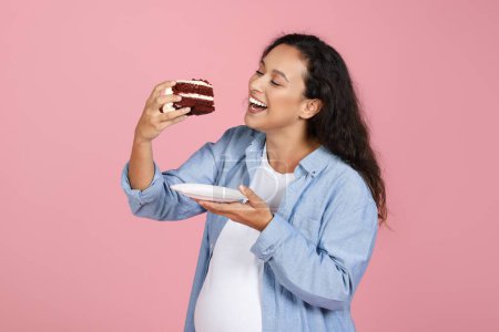 Foto de Embarazo Nutrición, Dieta. Emocionada mujer embarazada comiendo tarta de terciopelo rojo malsana Postre de pie sobre fondo de estudio rosa, ansia de carbohidratos - Imagen libre de derechos