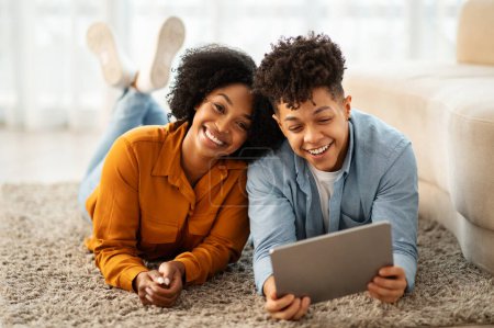 Téléchargez les photos : Allongé sur un tapis shaggy, un jeune couple afro-américain joyeux partage une tablette, leurs rires et leur joie remplissant la pièce pendant qu'ils profitent d'un moment décontracté dans une maison lumineuse et moderne - en image libre de droit