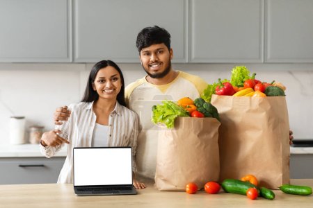 Smiling millennial eastern couple order grocery online, standing next to table at kitchen, holding paper bag full of fresh organic fruits and vegetables, point at laptop with blank screen copy space