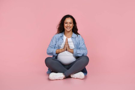 Foto de Mujer embarazada hermosa pacífica vistiendo ropa casual sentada en el suelo, cogida de la mano en gesto namaste, meditando sobre el fondo rosa. Alivio del estrés durante el embarazo - Imagen libre de derechos