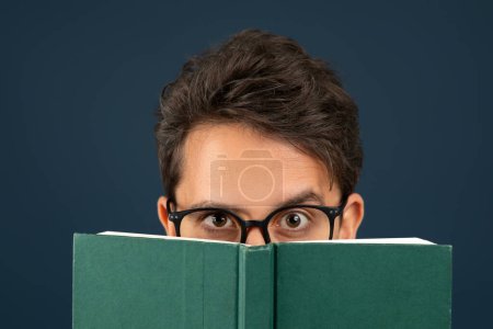 Foto de Joven con anteojos mirando por encima del libro verde, hombre nerd mostrando curiosidad e interés, de pie aislado en el fondo del estudio azul oscuro, disfrutando de la lectura, primer plano - Imagen libre de derechos