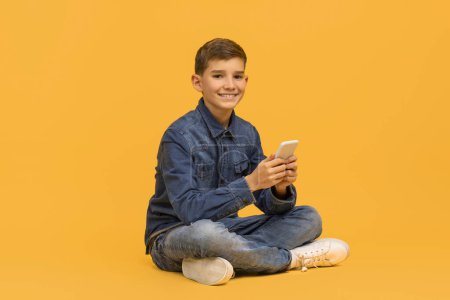 Photo for Smiling teen boy with smartphone in hands sitting cross-legged on vivid yellow background, happy male kid engrossed in using his mobile phone, depicting connectivity and casual technology use - Royalty Free Image