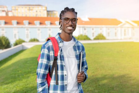 Photo for African American Student Guy With Backpack And Laptop Computer Stands Outside University Building, Posing Outdoor And Smiling To Camera. E-Learning And Educational Gadgets - Royalty Free Image