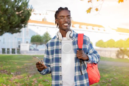 Téléchargez les photos : Joyeux étudiant noir de l'université avec sac à dos et écouteurs en utilisant un smartphone sur le campus à l'extérieur, incarnant l'éducation moderne et la technologie, naviguer dans les applications éducatives - en image libre de droit