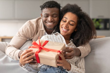 Téléchargez les photos : Romantique présent. Aimant mari noir présentant boîte cadeau à la femme à la maison à l'intérieur, célébrant un anniversaire de relation ensemble, étreignant sur le canapé dans le salon. Offre Saint-Valentin - en image libre de droit