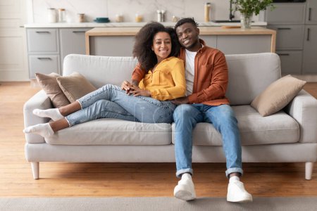 Téléchargez les photos : Maison Comfort. Heureux jeune couple de la famille noire se refroidissant sur le canapé dans le salon moderne, souriant à la caméra tout en étreignant. Conjoints se reposant ensemble le week-end, profiter du temps libre - en image libre de droit