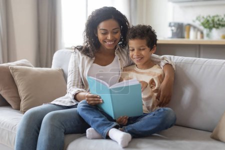 Photo for Family leisure activity. Young black mother and little son reading book together, happy african american mom and preteen male child sitting on couch, enjoying spending free time at home - Royalty Free Image